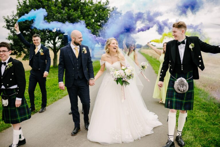 Bride and groom walking down the Bunny Hill venue lane with coloured smoke bombs and groomsmen in kilts – a summer barn wedding in Yorkshire