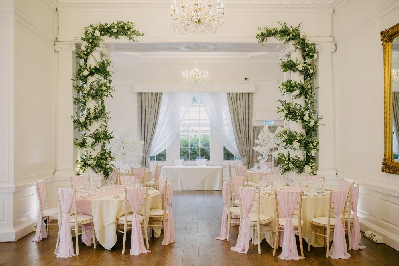 The ceremony room at Bawtry Hall set for the wedding, with a floral arch in front of the tall Georgian windows and blush pink chair sashes