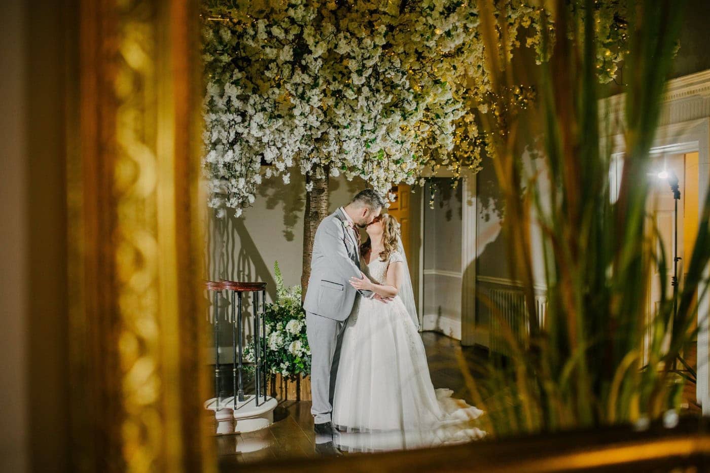 The bride and groom by the sweeping staircase covered in blossom at Bawtry Hall, reflected in a gold mirror at dusk