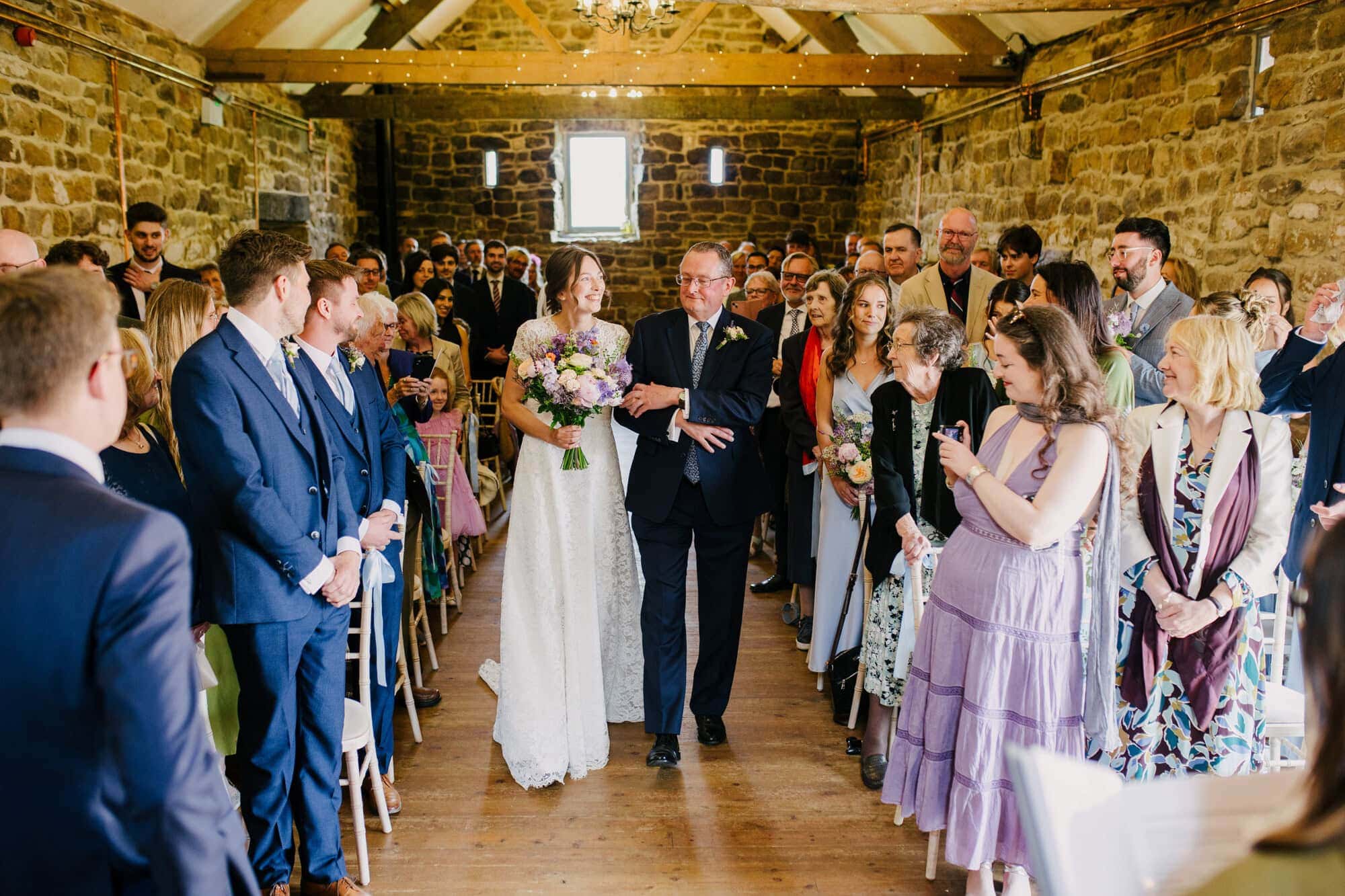 Ellie walking down the aisle with her dad in the stone barn at Danby Castle as George waits at the front
